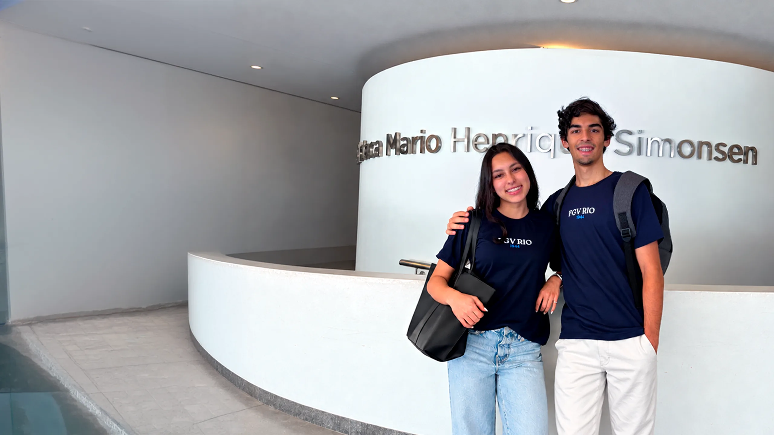 Dois alunos da FGV Rio sorrindo, de camiseta azul-marinho da FGV, no hall da Biblioteca Mario Henrique Simonsen