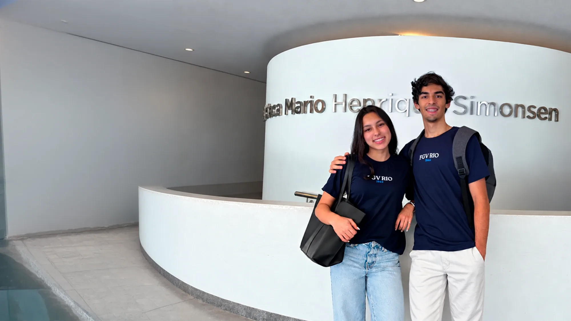 Dois alunos da FGV Rio sorrindo, de camiseta azul-marinho da FGV, no hall da Biblioteca Mario Henrique Simonsen